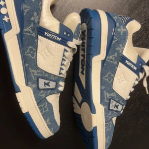 Louis Vuitton blå-vita sneakers  - Säljer ett par Louis Vuitton sneakers i blått och vitt med klassiskt monogrammönster. Skorna har en chunky siluett, snörning och platt sula. Materialmix av denim och präglat skinn, med tydliga LV-loggor på tungan och sidan. Perfekt för dig som gillar exklusiv streetstyle. Använd fåtal gånger.
