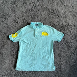 Ralph Lauren piké - Turkos kortärmad polo t-shirt från Ralph Lauren med klassisk krage och tre knappar framtill. Stor gul broderad logga på bröstet och ett gult emblem på ena ärmen. Tillverkad i mjuk bomull, perfekt för chill dagar. Snygg passform och stilren design. Storlek L 14-16 år vilket bör passa som XS men de måtten. :)