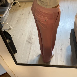 Rosa mjukisbyxor Juicy Couture - Säljer ett par rosa mjukisbyxor från Juicy Couture med bred midja och snörning framtill. Byxorna har raka ben, fickor med lock och silverfärgade knappar bak. Mjukt och skönt material som ger en chill vibe, perfekt för häng eller mysiga dagar. Stretchiga i midjan💕💕