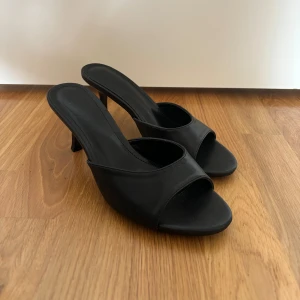 Svarta klackade sandaler i skinn - Snygga svarta sandaler med öppen tå och låg klack. De har en minimalistisk design med bred rem över foten och är tillverkade i slätt skinn. Perfekta för dig som gillar stilrena och trendiga skor med en elegant touch. Aldrig använda och helt nya! 