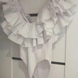 Vit body med volanger - Supersnygg vit bodysuit med breda volanger runt axlarna och djup v-ringning. 