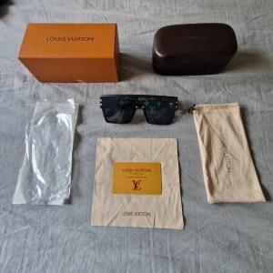 Blå Louis Vuitton solglasögon monogram - Säljer ett par blå solglasögon från Louis Vuitton med rektangulär form och breda skalmar. Glasen har LV monogram-mönster i blått och grönt, vilket ger en riktigt lyxig vibe. Kommer med originalfodral, ask, putsduk och certifikat. Perfekt för dig som vill sticka ut.