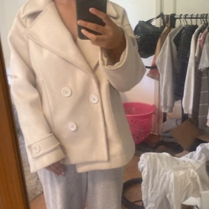 Vit skepparkavaj med stora knappar - Säljer en vit skepparkavaj med breda slag och dubbelknäppning framtill. Jackan har stora vita knappar både på framsidan och vid ärmsluten, samt en loose passform som ger en chill vibe. Materialet känns som ull och är mjukt och varmt, perfekt för kyliga dagar.