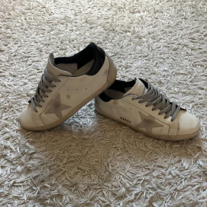 GOLDEN GOOSE- vit blåa sneakers med originella tags/ dustbag - Säljer ett par snygga Golden Goose sneakers i vitt skinn med grå mockastjärna på sidan och grå snörning. Skorna har svart häl och GGDB-logga i metall. Rund tå och platt sula ger en klassisk vibe. Perfekta för dig som gillar streetstyle och coola detaljer. Oanvända och helt nya med alla originella tags och kvitteringar. Dustbag medföljer! 