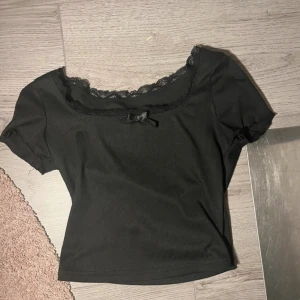 Svart croppad topp med spets och rosett - Supersöt svart croppad topp med korta ärmar, spetskant runt halsen och en liten rosett framtill. Ribbad struktur i tyget ger en extra touch. Perfekt för dig som gillar en lite gullig men ändå stilren vibe.