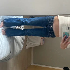 Blå bootcut jeans - Säljer ett par blå jeans med bootcut passform och låg midja. Jeansen har tydliga vita kontrastsömmar, dekorativa ficklock med knappar och klassisk logga på bakfickan. Materialet är jeans med stretch för skön passform.
