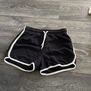 Svarta sportshorts med vit kant - Svarta sportiga shorts från C med vit kant och snörning i midjan. Modellen har rundade ben och elastisk midja för skön passform. Perfekta för träning eller chill dagar. Materialet känns mjukt och lätt, troligen bomull eller polyester.