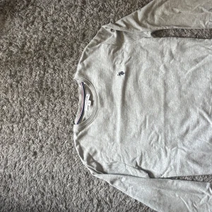 Grå långärmad tröja från U.S. Polo Assn. - Snygg grå långärmad tröja från U.S. Polo Assn. med klassisk rund hals och diskret broderad logga på bröstet. Tröjan har en stilren look med marinblå och röda detaljer i halsen. 