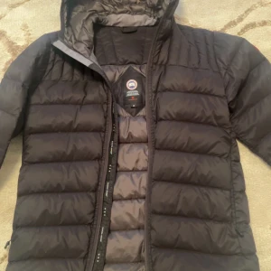 Svart dunjacka från Canada Goose - Snygg svart dunjacka från Canada Goose med huva och klassisk logga på ärmen. Jackan har quiltad design, dragkedja framtill och är perfekt för kalla dagar. Materialet är polyester och dun, vilket gör den både lätt och varm. Passformen är normal och jackan har långa ärmar.