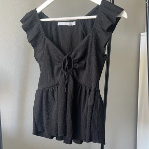 Svart blus med volang och knyt XS - Supersöt svart blus från 157 i storlek XS. Blusen har volangdetaljer vid axlarna, knyt framtill och en lätt rynkad struktur i tyget. Modellen är lite peplum med en luftig passform och ärmlös design. Perfekt för dig som gillar stilrena och trendiga toppar.