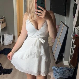 Vit klänning med volanger - Säljer en söt vit klänning med volanger och knyt i midjan. Klänningen har v-ringad hals, är ärmlös och har ett diskret prickigt mönster. Perfekt för sommarens alla tillfällen, superfin och lätt att bära. Stl s men passar även m