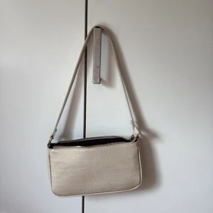 Beige axelväska med croco-mönster - Snygg beige axelväska med croco-mönster och stilren form. Väskan har ett huvudfack med dragkedja, innerficka med zip och flera små fack för dina essentials. Perfekt storlek  för mobilen, plånboken och lite smink. Materialet är syntet med glansig finish.