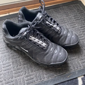 Nike tns - Säljer ett par svarta Nike sneakers med coolt vågigt mönster och silvriga detaljer på snörningen. Skorna har en platt sula och är tillverkade i mesh och syntetmaterial för skön komfort. Perfekta för dig som gillar sportig stil och vill ha något som sticker ut.