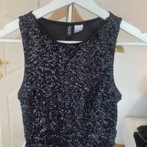 Svart glittrig paljettopp från H&M - Svart ärmlös topp från H&M Divided, täckt med glittrande paljetter på framsidan som ger en riktigt festlig vibe. Baksidan är i mjuk, slät polyester. Rund halsringning och croppad passform som sitter snyggt. 