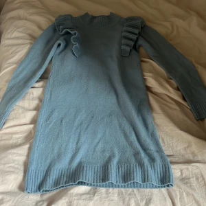 Blå stickad klänning - Snygg ljusblå stickad klänning som går ner till knäna  med långa ärmar och coola volangdetaljer på axlarna. Klänningen har ribbade muddar vid hals, ärmslut och nederkant. Perfekt för dig som gillar en chill men ändå unik look.