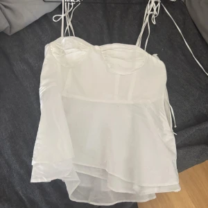Vit linnetopp med snörning - Säljer en vit omlott linnetopp med tunna axelband och snörning. Toppen har en lätt glansig yta och volangdetaljer nedtill. Bysten är formad och ryggen har smock för extra komfort. Perfekt för dig som gillar en luftig och feminin stil.