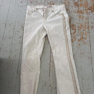 Vita jeansbyxor från Mos Mosh - Snygga vita jeansbyxor från Mos Mosh med raka ben och beige dekorativa ränder längs sidorna. Klassisk femficksmodell med blommigt tyg på insidan av linningen. Perfekt för dig som vill ha en fräsch och stilren look.