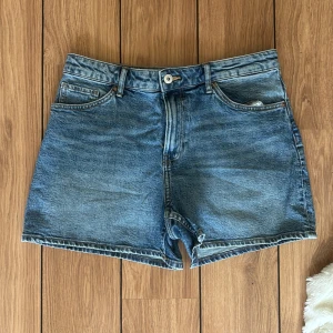 Blå jeansshorts från H&M, strl 40 - Snygga blå jeansshorts från H&M i klassisk femficksmodell med hög midja och normal passform. Shortsen har bälteshällor, knapp och dragkedja framtill samt slitstarka sömmar. Perfekta för sommarens alla äventyr!