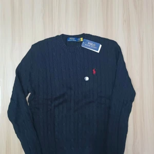 Mörkblå stickad tröja Polo Ralph Lauren - Snygg mörkblå stickad tröja från Polo Ralph Lauren med klassiskt kabelstickat mönster. Tröjan har rund hals och lång ärm, samt den ikoniska röda Polo-loggan broderad på bröstet. Perfekt för dig som gillar stilrena och tidlösa plagg.