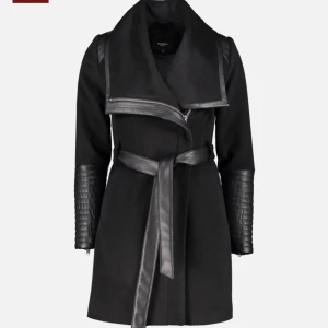 Svart kappa med skinn-detaljer - Lyft din outfit med denna eleganta svarta kappa som kombinerar klassisk design med en modern edge. Kappan är tillverkad i mjukt, följsamt material och har en smickrande skärning som markerar midjan med ett avtagbart skärp.  De snygga detaljerna i läder på axlar och ärmar ger jackan en exklusiv och trendig känsla, medan det stora slaget och asymmetriska dragkedjan skapar en sofistikerad silhuett.  ✔️ Färg: Svart ✔️ Material: Mjuk textil med läder ✔️ Stängning: Dragkedja + skärp i midjan ✔️ Passform: 