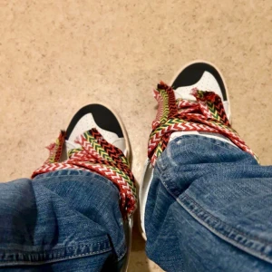 Lanvin sneakers med färgglada snören - Storlek 41 men dem passar i 41.5 Unika Lanvin sneakers i vitt skinn med svarta detaljer och breda, färgglada snören i rött, gult, grönt och vitt zigzagmönster. Snygg kontrast mellan den klassiska siluetten och de iögonfallande snörena. Perfekta för dig som vill sticka ut med din stil.