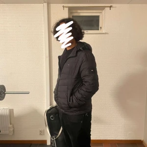 Svart pufferjacka Tommy Hilfiger - Svart pufferjacka från Tommy Hilfiger med huva och dragkedja framtill. Jackan har quiltad design och logotyp på ärmen. Perfekt för kalla dagar och har en clean, modern look som funkar till streetwear. Materialet är polyester och jackan är långärmad. Pris kan diskuteras privat.