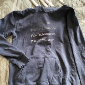 Mörkblå hoodie från Peak Performance - Säljer en mörkblå hoodie från Peak Performance med tryckt logga i olika nyanser på ryggen. Tröjan har huva, magficka och ribbade muddar vid ärmslut och nederkant. Perfekt för chill eller häng med kompisar.