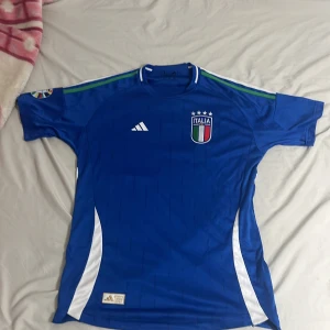 Italien Chiesa 14 blå fotbollströja Adidas - Säljer en officiell Italien fotbollströja från Adidas med Chiesa och nummer 14 på ryggen. Tröjan är blå med vita detaljer, har korta ärmar och broderat landslagsmärke på bröstet. Materialet är lätt och andas, perfekt för match eller träning.