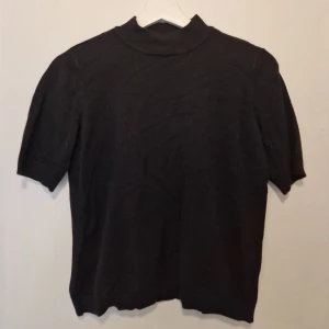 Svart polotröja med kort ärm - Svart polotröja med enkel design med en liten metallring i nacken som detalj. Perfekt för dig som gillar stilrena och minimalistiska plagg.