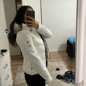 Vit dunjacka med huva från moncler - Snygg vit dunjacka från Moncler med huva och dragkedja framtill. Jackan har quiltad kropp, släta ärmar och en patch med logga på ena ärmen. Perfekt för kalla dagar och har en clean, modern look. Storlek S och i nyskick, säljs pga för liten storlek (aldrig använd). Pris kan diskuteras vid snabb affär.