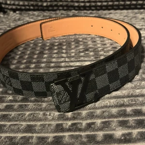 Louis Vuitton rutig svart skinnbälte - Säljer ett Louis Vuitton bälte i svart och grått med klassiskt rutigt Damier-mönster och en stor LV-logga i metall på spännet. Bältet är gjort i äkta skinn med beige insida och har en stilren, modern vibe som lyfter vilken outfit som helst.