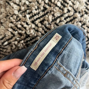 Levi's 725 High Rise Bootcut Jeans - Säljer ett par ljusblå Levi's 725 High Rise Bootcut jeans i storlek 25. De har klassisk femficksdesign, mellan hög midja och bootcut-ben som ger en snygg siluett. Jeansen är tillverkade i mjukt denim med Levi's ikoniska läderpatch och röda logotagg bak.