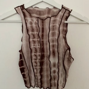 Brun och beige ärmlös linnetopp - Säljer en tunn ärmlös linnetopp i beige och bruna toner med ett randigt mönster. Toppen har vågiga kanter och synliga sömmar som ger en edgy vibe. Materialet är lätt och semi-transparent, perfekt för att styla lager på lager.