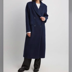 Lång mörkblå kappa i ullblandning från NA-KD - Oversized mörkblå kappa från NA-KD. Lång modell med raka ärmar och fickor framtill. Tillverkad i 50% ullblandning. Modell: 175 cm och har strl EU 36. NYPRIS:1699 kr
