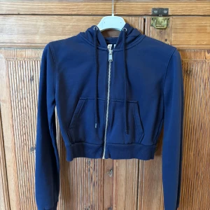 Mörkblå croppad hoodie från H&M Divided - Säljer en mörkblå croppad hoodie från H&M Divided. Tröjan har dragkedja framtill, huva med snören och två fickor. Perfekt för en avslappnad stil och enkel att matcha med jeans eller kjol. Storlek S.