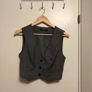 Grå väst från Gina Tricot - Stilren kroppad grå väst från Gina Tricot med tre knappar framtill och två dekorativa fickor. Klassisk passform med v-ringning, perfekt att styla över en skjorta eller t-shirt. Gjord i ett slätt tyg som ger en dressad look.