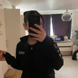  track jacket från Hoodrich - Svart track jacket från Hoodrich med vit logga längs ärmarna och på bröstet. Jackan har dragkedja framtill och hög krage. Sportig och stilren design som passar perfekt till streetwear.