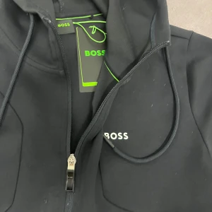 Svart hoodiejacka från BOSS - Svart hoodiejacka från BOSS med dragkedja framtill och vita detaljer vid fickorna. Jackan har huva med dragsko och BOSS-logga på bröstet. Perfekt för en sportig och stilren look.