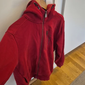 Röd hoodie från Sail Racing som bara har änvänd 7 gånger  - Röd hoodie från Sail Racing med dragkedja framtill och på bröstfickan. Tröjan har huva med snörning och logga på bröstet. Mjuk bomullskänsla och normal passform, perfekt för en avslappnad stil. Storlek L