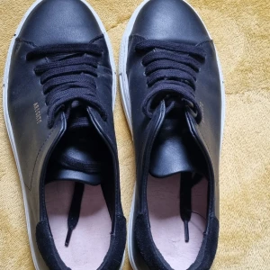 Svarta sneakers från Arigato - Snygga svarta sneakers från Arigato med klassisk design och vita sulor. Skorna har svarta snören och är tillverkade i skinn. Perfekta för en stilren och enkel look. Märket syns i guld på sidan av skon. Behövs nya innersulor.