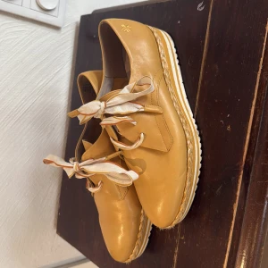 Gula läderskor med rosett från Marco Tozzi - Säljer ett par snygga gula läderskor från Marco Tozzi med breda, mönstrade snören som knyts i rosett. Skorna har rund tå, platt sula och dekorativ söm runt sulan. Perfekta för dig som vill sticka ut med färg och stil.