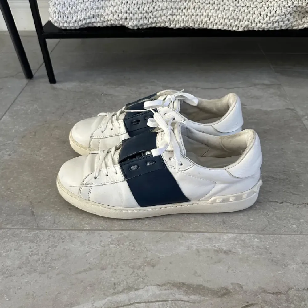 Byten går bättre än cash ‼️Säljer ett par snygga Valentino Open sneakers i vitt skinn med bred marinblå rem över mitten. Skorna har klassisk snörning, rund tå och platt sula med ikoniska nitar baktill på sulan. Perfekta för dig som gillar stilrena och exklusiva sneakers.. Kengät.