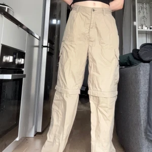Beiga cargopants med avtagbara ben - OBS!! ALLA MINA PLAGG GRATIS VID UPPHÄTMNING I RÅCKSTA!!! Säljer ett par beiga cargopants med dragkedja vid knäna så de kan göras om till shorts. Byxorna har flera fickor på sidorna och en avslappnad passform. Grymma på sommardagar med omväxlande väder. De är tunna och luftiga. Jag klämmer åt midjan på bilderna, de är ganska stora men funkar med ett bälte på mig som är storlek S annars. 