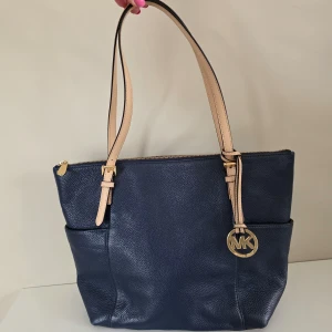 Mörkblå axelväska från Michael Kors - Mörkblå axelväska från Michael Kors med beige axelremmar och guldfärgade detaljer. Väskan har dragkedja upptill, två ytterfickor och ett stort innerfack med logotypmönstrat foder. 