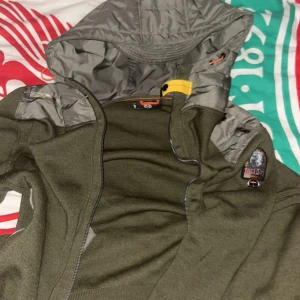 Olivgrön jacka med huva från Parajumpers - Säljer en olivgrön jacka från Parajumpers med grå vadderad huva och detaljer. Jackan har dragkedja och en gul rem vid halsen. Märkespatch på ärmen. Perfekt för dig som vill ha en snygg och funktionell jacka.