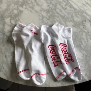Vita strumpor med röd Coca-Cola text 2 par - Säljer 2 par vita strumpor med röda detaljer och Coca-Cola logga. Strumporna har röda ränder och text längs sidan. Perfekta för dig som gillar klassiska och snygga strumpor med retrovibbar. Aldrig använda!  strl 43-46