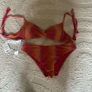 Glittrig calzedonia bikini i orange och rosa  - Säljer en snygg bikini med glittrigt tyg i orange och rosa nyanser. Överdelen har omlott-design och knytband både i ryggen och nacken. Bikinitrosan har en klassisk passform. Perfekt för stranden eller poolen!