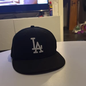 Svart LA keps från New Era 59FIFTY - Säljer en svart keps från New Era 59FIFTY med LA-logga broderad framtill i vitt. Klassisk flat peak och broderad New Era-logga på sidan. Perfekt för dig som gillar sportig stil.
