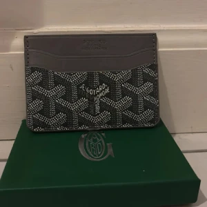 Grå korthållare - Säljer en stilren grå korthållare med det klassiska mönstret och logotypen framtill. Korthållaren har flera fack för kort och är tillverkad i skinn med detaljer i syntet. Perfekt för dig som vill ha en exklusiv accessoar.