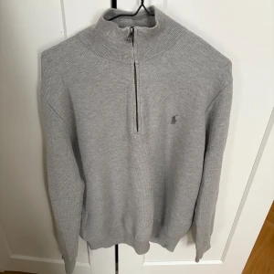 Grå stickad tröja med halv dragkedja från Polo Ralph Lauren - Säljer en stilren grå stickad tröja från Polo Ralph Lauren i storlek M. Tröjan har halv dragkedja vid halsen och diskret broderad logga på bröstet. Perfekt för lager-på-lager och passar till många olika outfits.